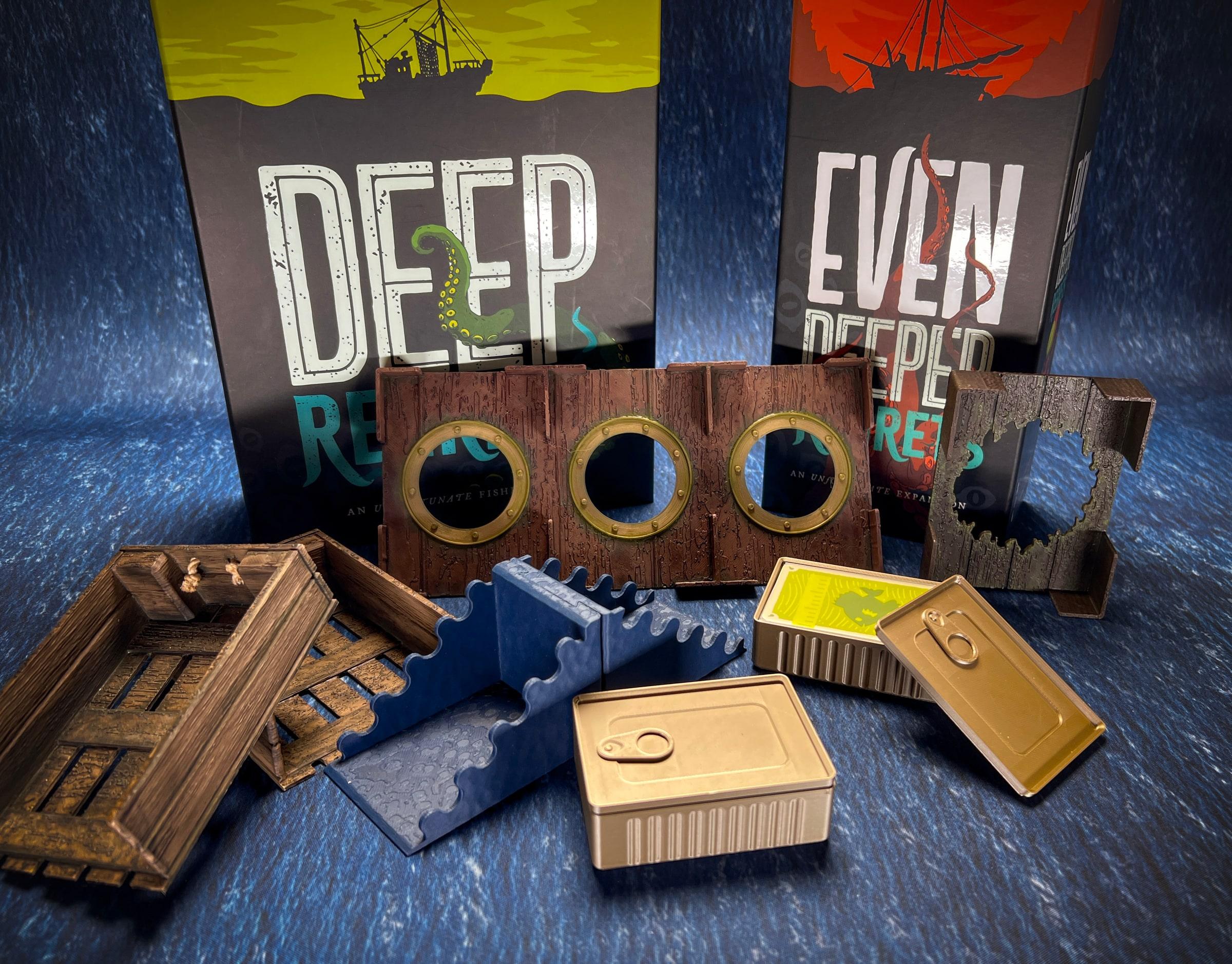 The Deep Regrets insert laid out on a blue sea mat: a porthole wall, wooden crates, a wave tray and sardine-tin card holders, with the game boxes behind.