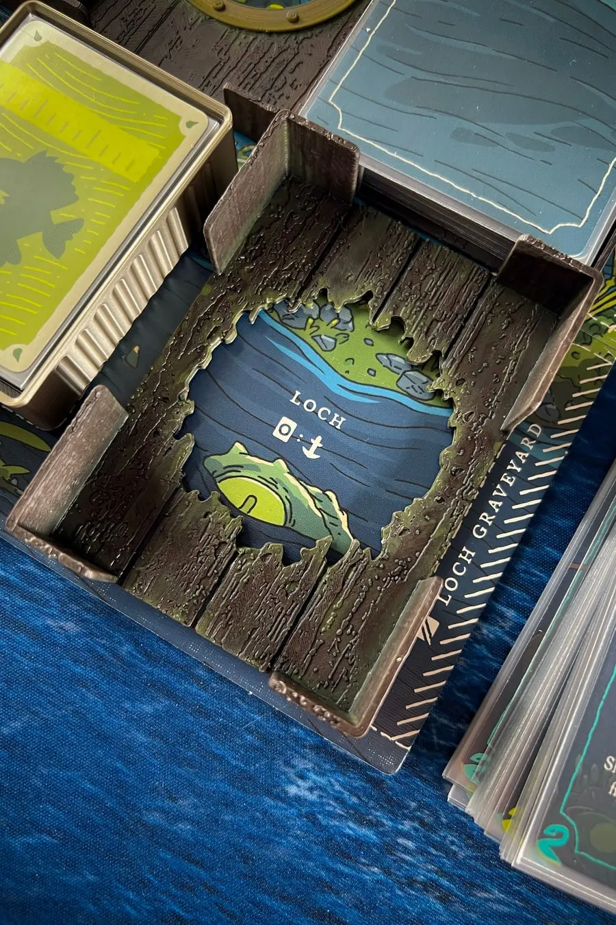 The printed Deep Regrets loch tray, a weathered wooden frame around the Loch card, on a blue sea mat.